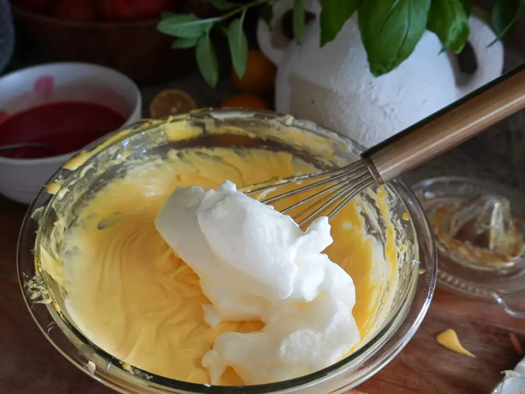 A glass bowl contains yellow batter and whisked egg whites being folded in with a whisk, with kitchen items in the background.