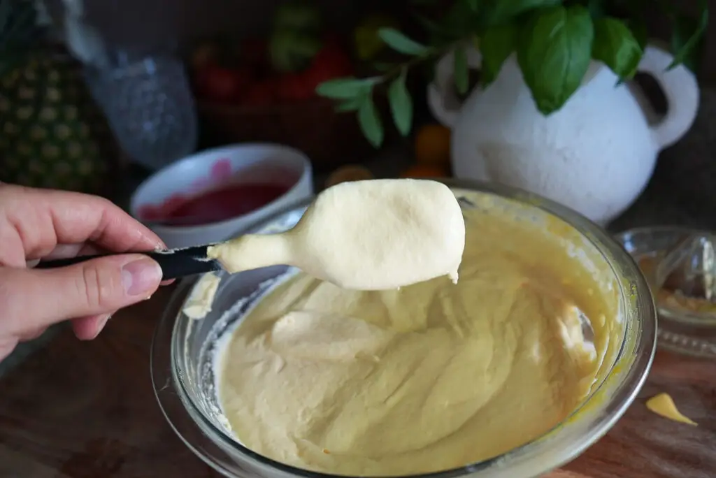 A hand holds a spatula with thick, yellow batter over a glass mixing bowl filled with the same batter on a kitchen counter.