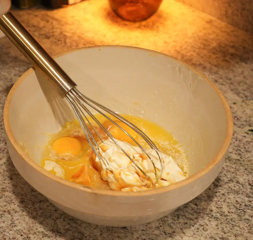 A metal whisk in a bowl containing raw eggs and sour cream.