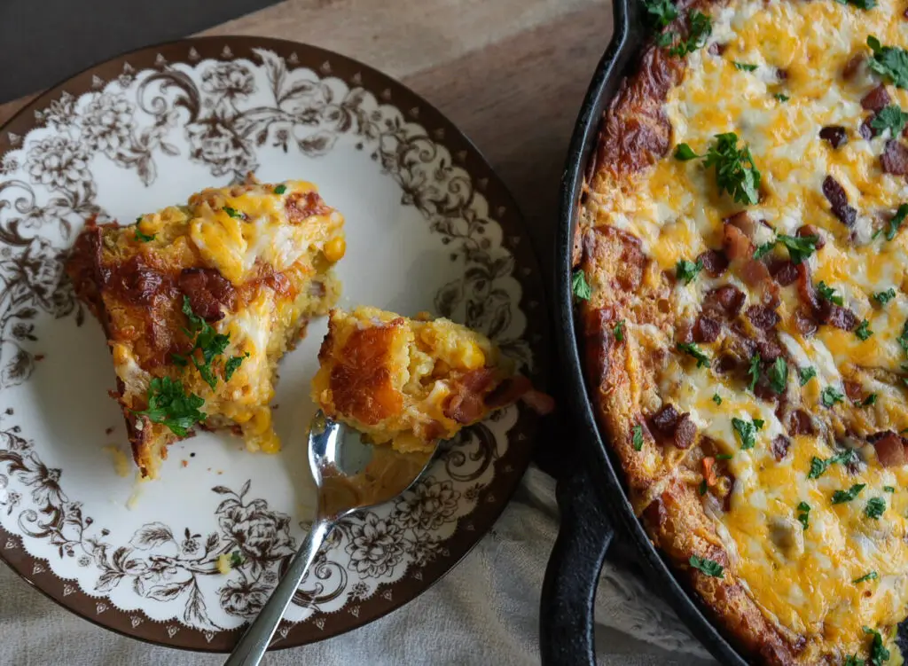 A serving of bacon and corn casserole on a decorative plate next to a cast-iron skillet containing the rest of the dish.