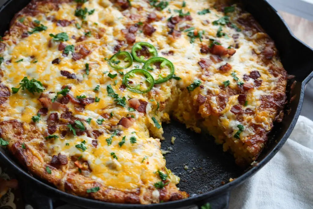 Cast iron skillet containing cornbread topped with melted cheese, bacon bits, jalapeño slices, and herbs, with a slice removed.