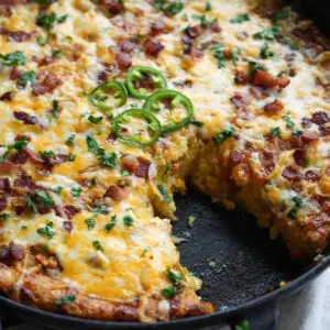 Cast iron skillet containing cornbread topped with melted cheese, bacon bits, jalapeño slices, and herbs, with a slice removed.