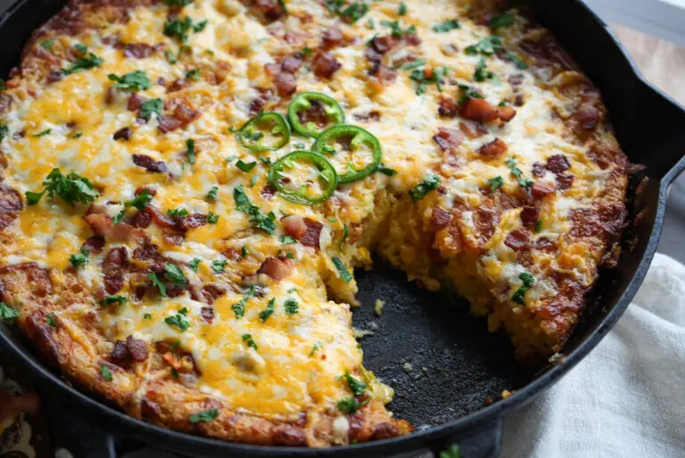 Cast iron skillet containing cornbread topped with melted cheese, bacon bits, jalapeño slices, and herbs, with a slice removed.