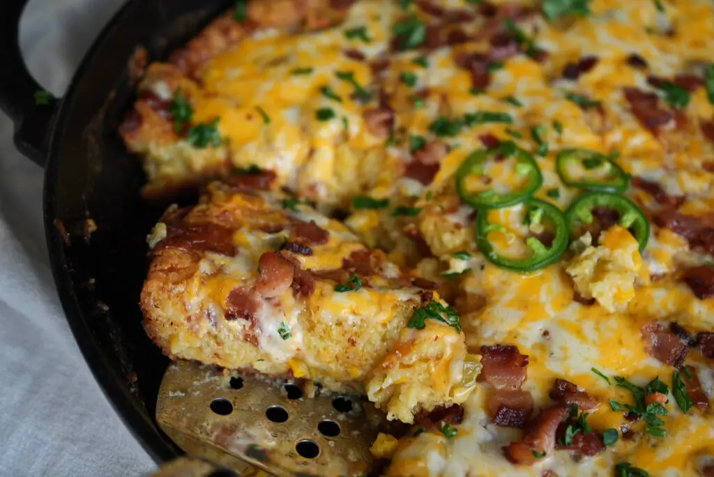 A slice of cheesy cornbread topped with bacon, jalapeño slices, and herbs being lifted from a cast-iron skillet with a metal spatula.