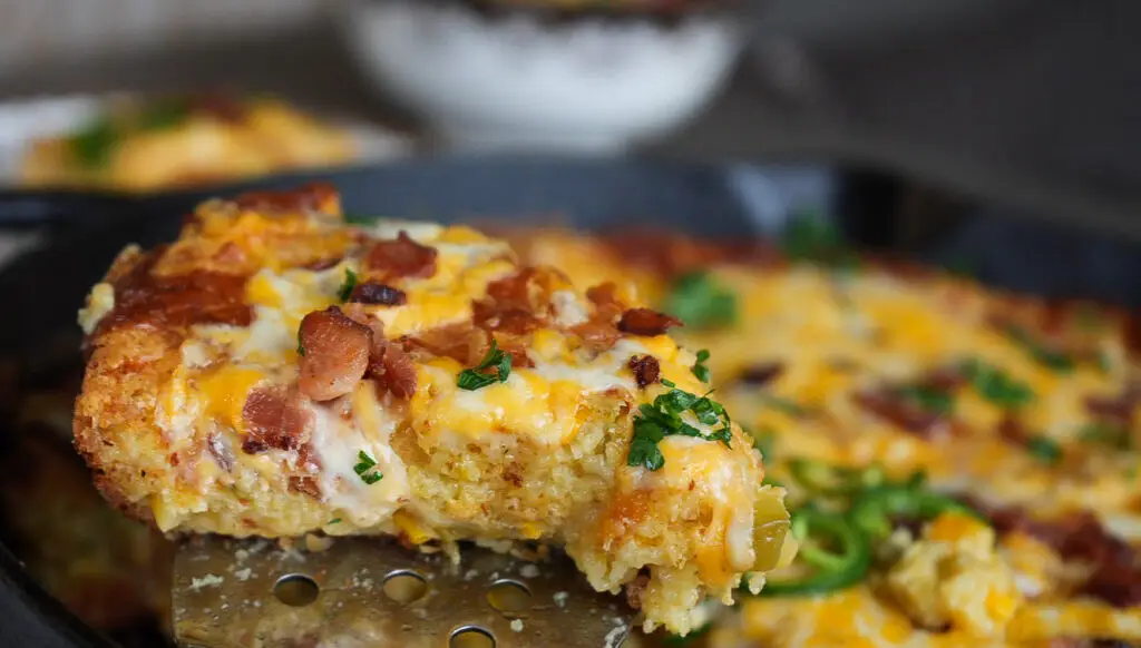 A slice of cheesy cornbread topped with bacon and herbs being lifted from a cast-iron skillet with a spatula.