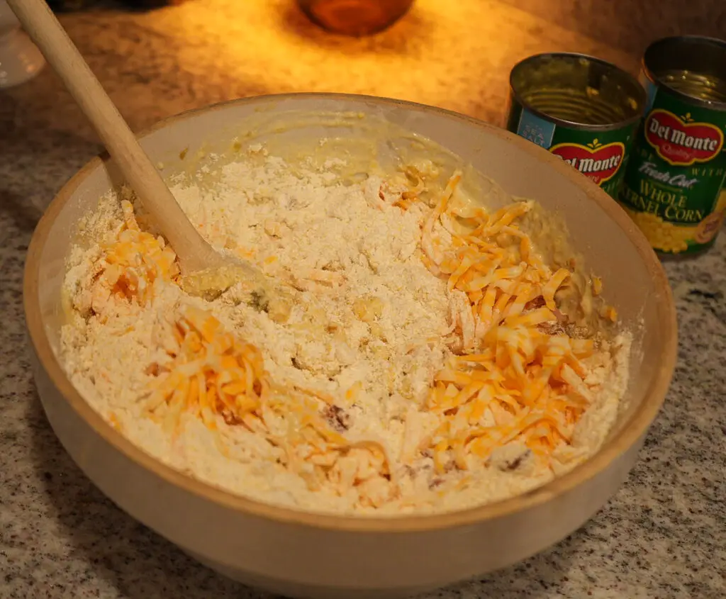 A wooden spoon stirs corn and shredded cheese into a bowl of dry baking mix, with two cans of Del Monte whole kernel corn nearby.