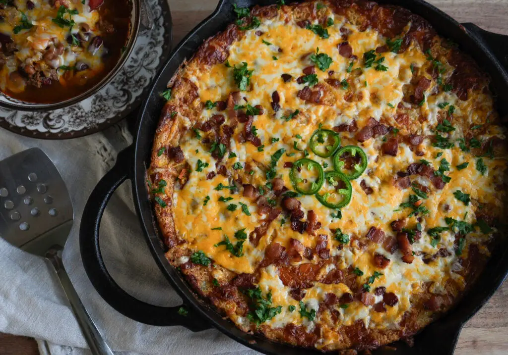 A cast iron skillet filled with a cheesy cornmeal dish topped with bacon, jalapeño slices, and herbs, next to a bowl of chili and a serving spatula.