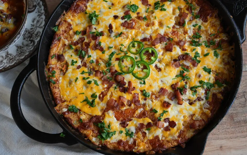 A cast iron skillet filled with a cheesy, bacon-topped casserole garnished with fresh jalapeño slices and herbs.