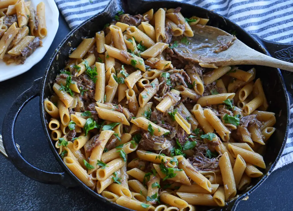 French onion soup pasta with shredded meat and herbs in a cast iron skillet, with a wooden spoon and a side plate of pasta.