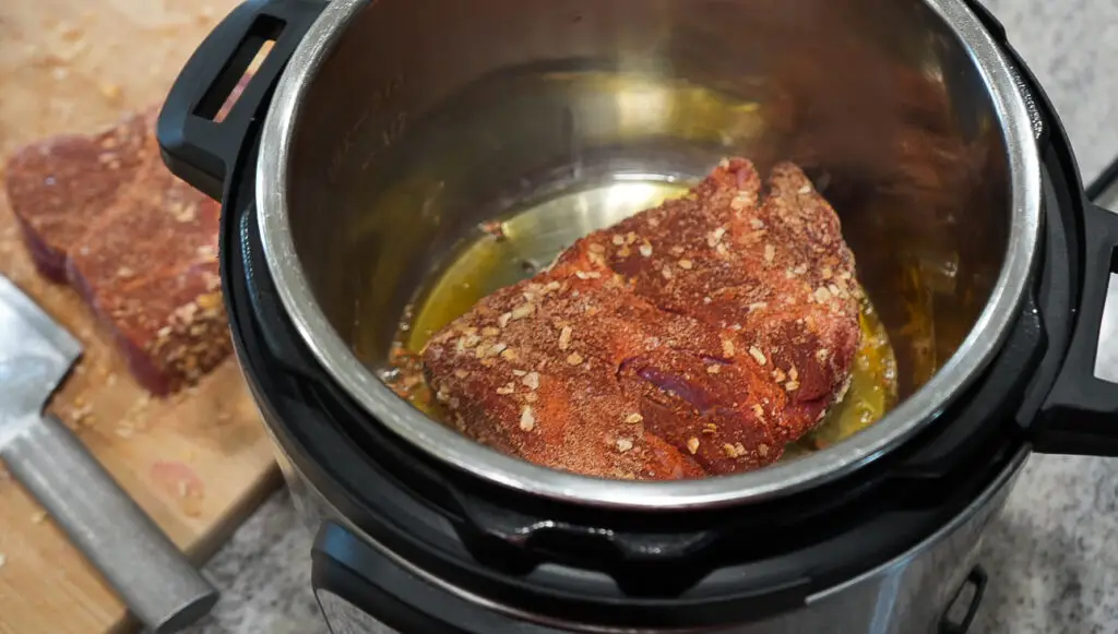 A seasoned piece of raw beef searing in an electric pressure cooker pot, with another piece of meat on a cutting board nearby.