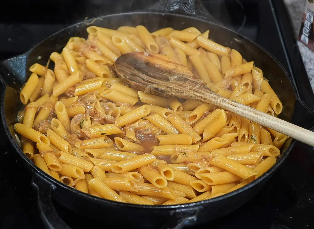 Penne pasta cooking in a cast-iron skillet with a wooden spoon.