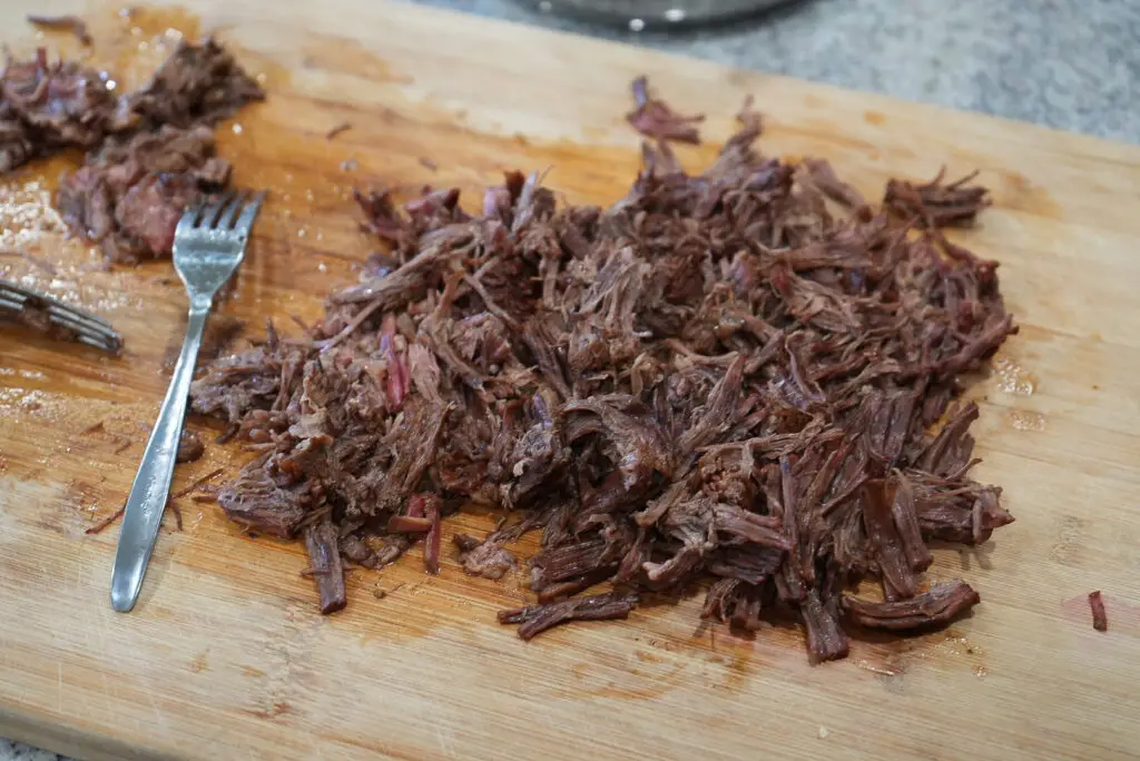 A pile of shredded cooked beef on a wooden cutting board with a metal fork.