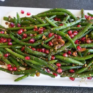 Cooked green beans topped with pomegranate seeds and chopped nuts on a white rectangular platter.