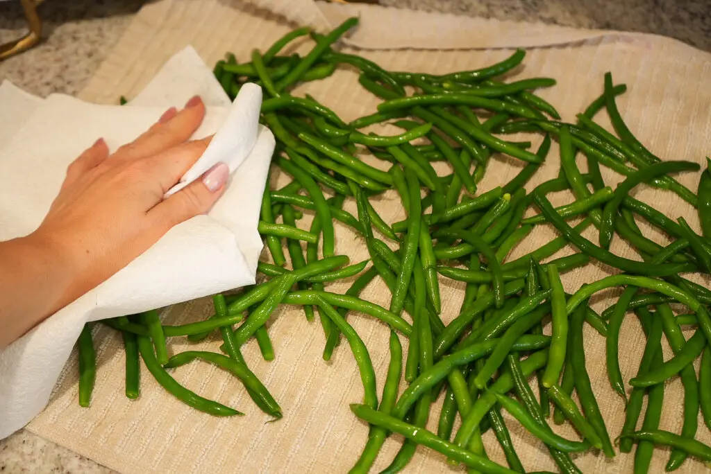 A hand uses a paper towel to dry green beans spread across a kitchen towel.
