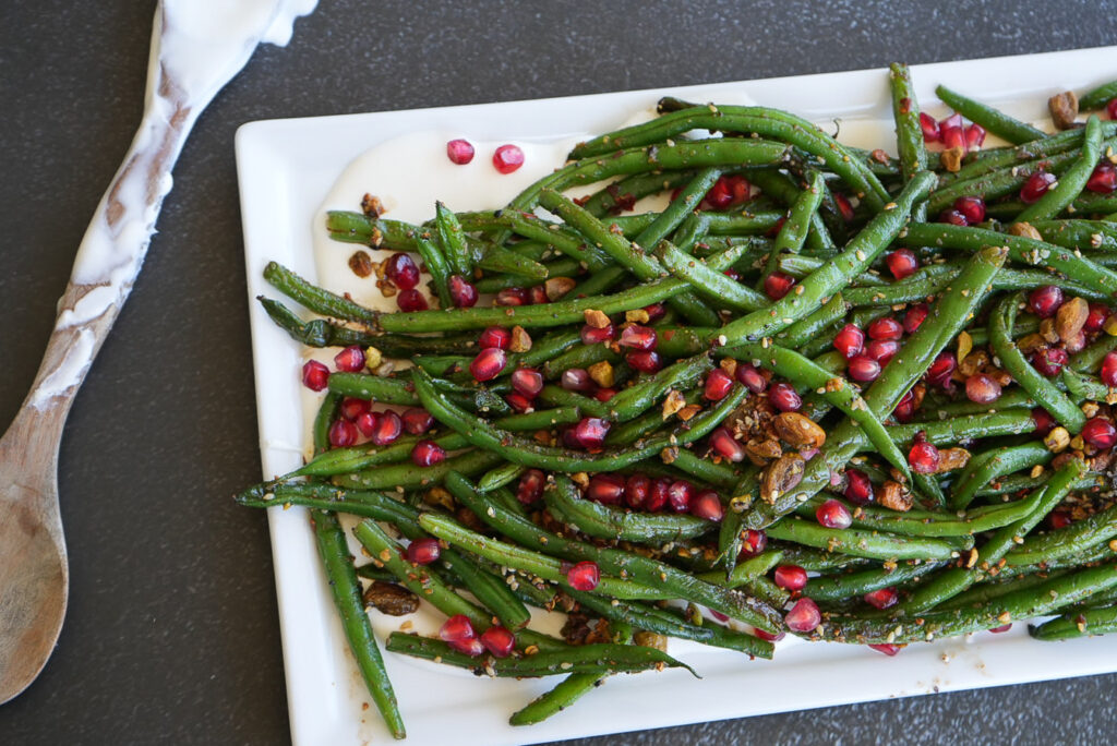 A white rectangular platter of cooked green beans topped with pomegranate seeds and chopped nuts, next to a wooden serving spoon.